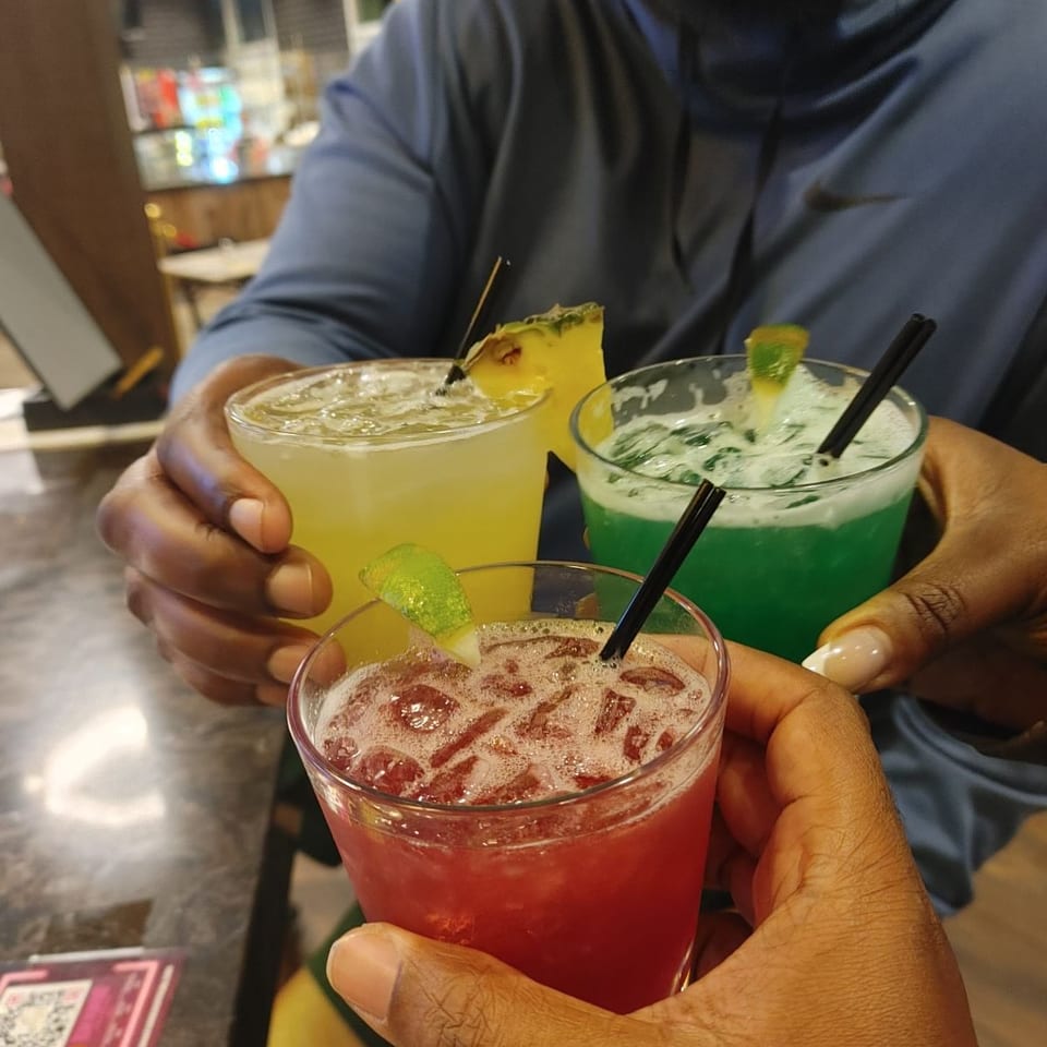 Three people raise colorful iced mocktails garnished with citrus and mint inside a brightly lit restaurant.