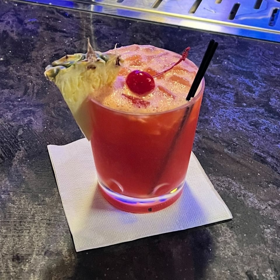 Red cocktail with foamy topping, cherry garnish, pineapple wedge, and black straw on a napkin-covered bar counter.