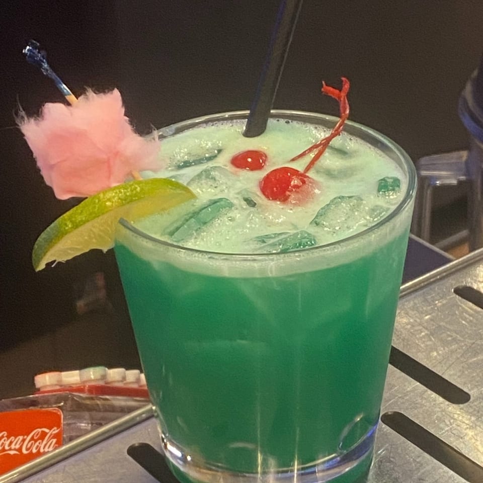 Bright green iced cocktail garnished with lime, cherries, a pink cotton-candy swizzle, and a straw on a bar counter.