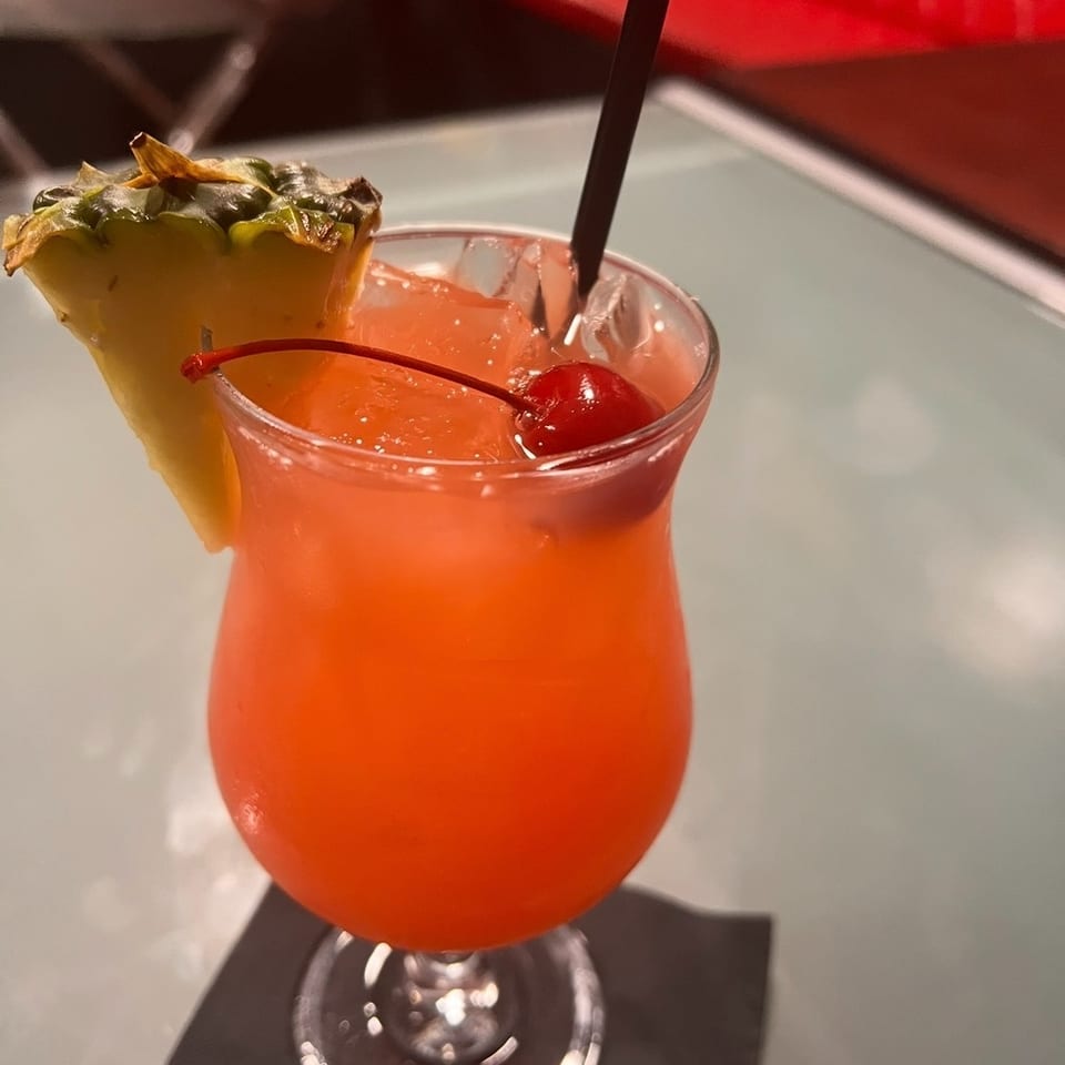 Bright red-orange cocktail in a stemmed glass, garnished with a pineapple wedge and cherry, with a straw on a restaurant table.